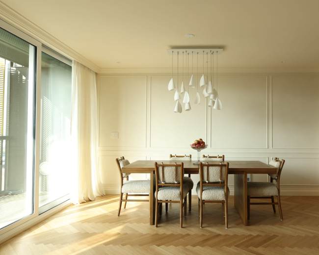 A dining area features a wooden table surrounded by four chairs and is illuminated by pendant lights, with large glass doors leading to an outside space.