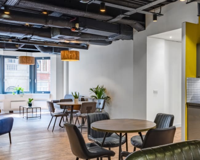 A modern office common area featuring round tables, black leather chairs, and a kitchenette with yellow accent walls.