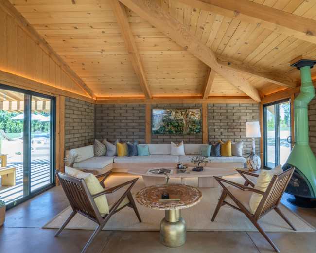 The image shows a modern living room with a wooden ceiling, a cozy sectional sofa, a coffee table, and a green fireplace, featuring large glass doors that open to an outdoor area.
