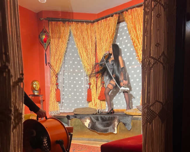 A person in lingerie poses on a platform in a room with orange walls and patterned curtains, while another person operates a large fan nearby.