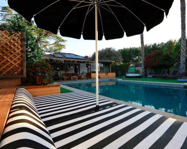 A black and white striped lounge chair and umbrella are positioned by a swimming pool surrounded by greenery and wooden decking.