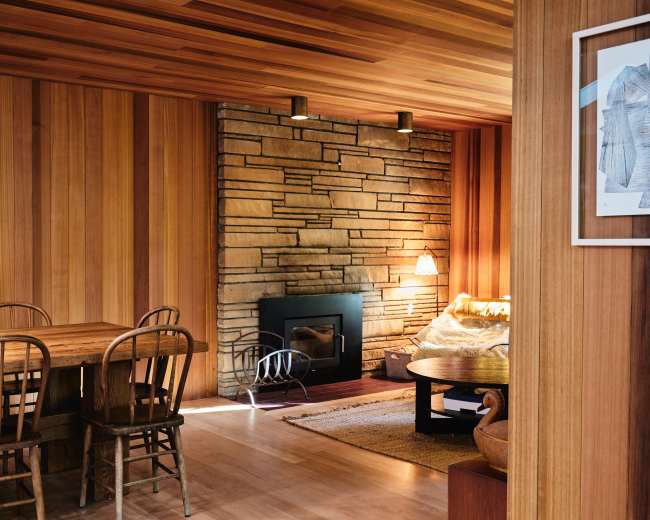 The image shows a cozy interior with wooden walls, a stone fireplace, and a dining table with chairs.