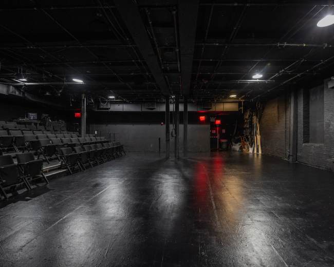 The image shows a black-box theater with empty rows of seats on one side and a dark stage area lit by overhead lights.