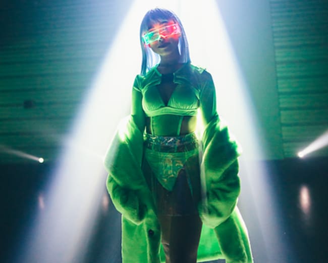 A person dressed in a brightly colored outfit stands illuminated by a beam of light on a stage.