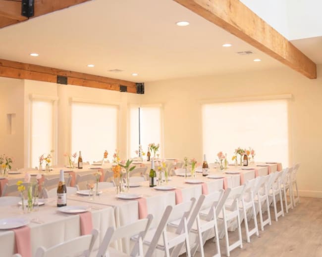 A spacious event room features long dining tables set with light tablecloths and floral centerpieces, illuminated by natural light from large windows.