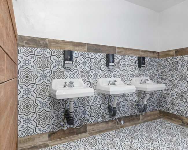 The image shows a bathroom with three white sinks mounted on a patterned tile wall, accompanied by wall-mounted soap dispensers.