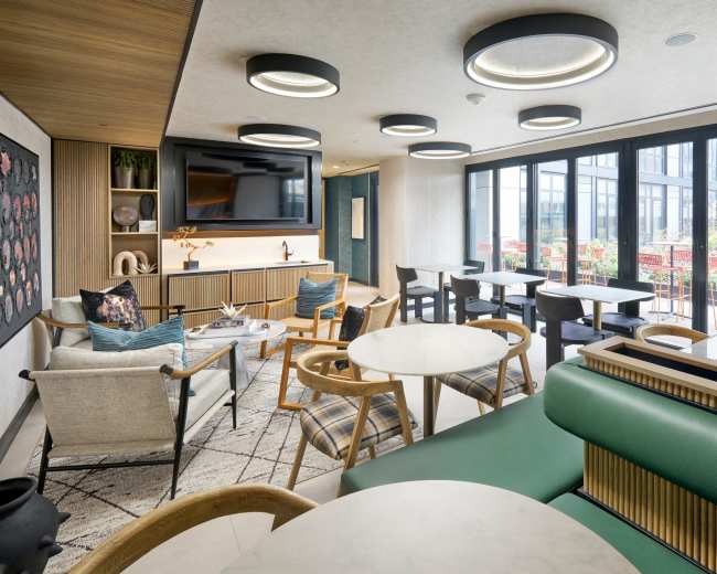 The image shows a modern lounge area with green seating, marble tables, and large windows overlooking an outdoor terrace.