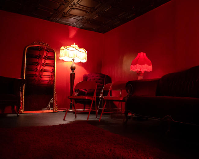 The scene features a red-lit room with a dark sofa, various chairs, and two lamps, one of which is a standing lamp and the other a table lamp.
