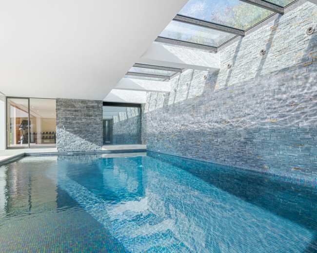The image shows an indoor swimming pool surrounded by walls made of stone and large glass panels letting in natural light.