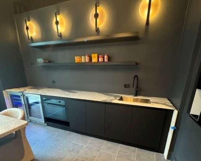 The image shows a modern kitchen with dark cabinetry, a light-colored countertop, and minimalistic shelves illuminated by circular lights.