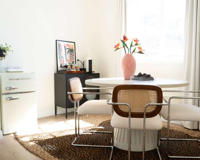 A round dining table surrounded by four chairs sits in a bright room, adorned with a vase of flowers and framed artwork, next to a vintage refrigerator and a textured rug.