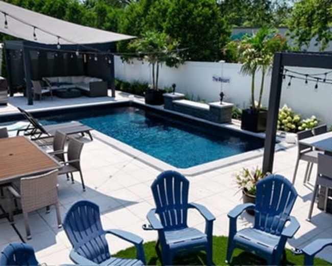 The image shows a landscaped backyard with a swimming pool, lounge chairs, outdoor seating, and a bar area under shaded structures.