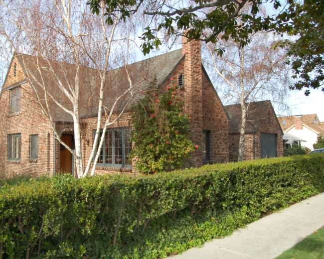 A brick house with a steep roof is surrounded by a neatly trimmed hedge and several birch trees.