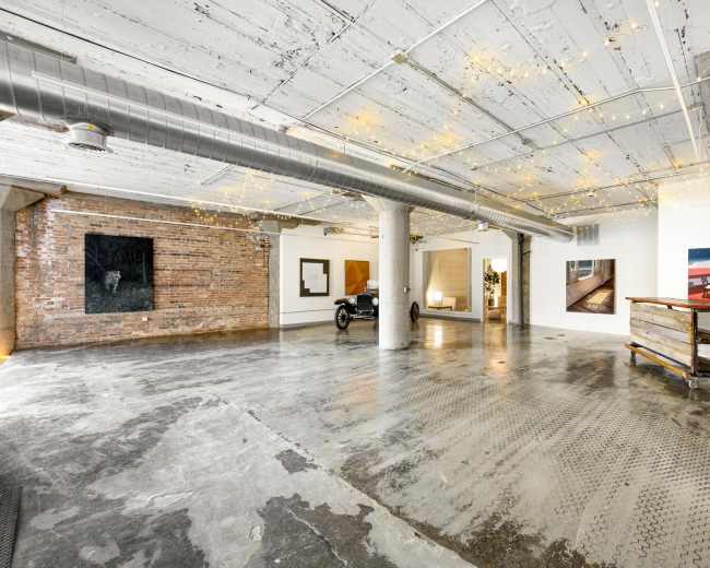 The image depicts a spacious industrial-style room with exposed brick walls, polished concrete flooring, and various artworks displayed on the walls.