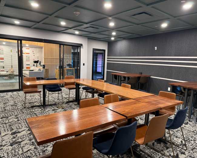 The image shows a modern meeting room with several wooden tables arranged in a U-shape and dark blue and black chairs, with a glass door leading to another area.