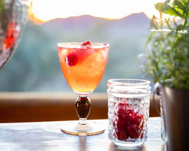 A cocktail with ice and raspberries is placed on a wooden surface with a blurred outdoor sunset in the background.