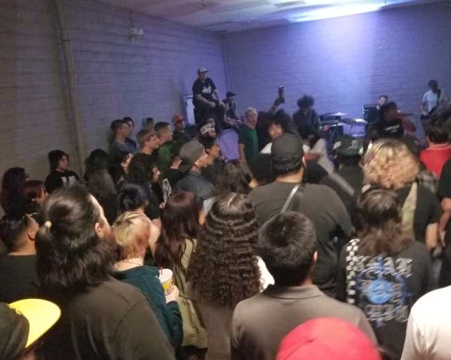 A crowd of people is gathered in a dimly lit room, watching a live band perform on stage.