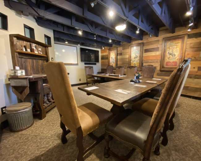 A conference room features a large wooden table surrounded by six chairs, with patterned walls and framed artwork in the background.