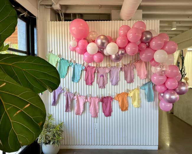 The scene features a wall adorned with pastel-colored baby clothes hanging from a string, complemented by a cluster of pink and white balloons.