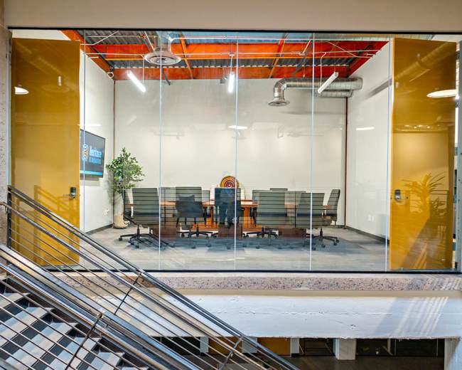 The image shows a glass-walled conference room with a long table and several chairs, surrounded by a staircase and a potted plant.