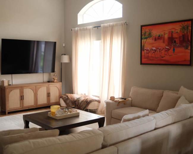The image depicts a well-lit living room featuring a large television mounted on the wall, a wooden entertainment unit, and a comfortable beige sofa set across from a coffee table.