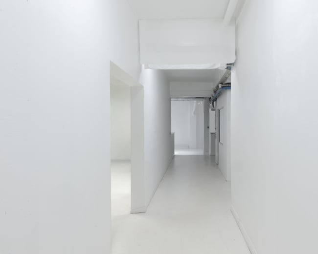 The image shows a long, narrow white hallway with doorways on either side and a bright, well-lit atmosphere.