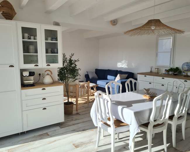 The image shows a bright, modern kitchen and living area featuring a white table with chairs, a sleek kitchen cabinet, and a cozy seating area with a blue sofa.