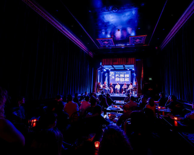 A small audience watches a panel discussion on stage in a dimly lit venue with blue lighting.