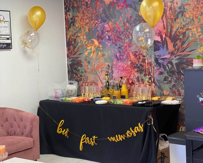 A party setup features a decorated table with snacks, drinks, and balloons against a vibrant floral backdrop.