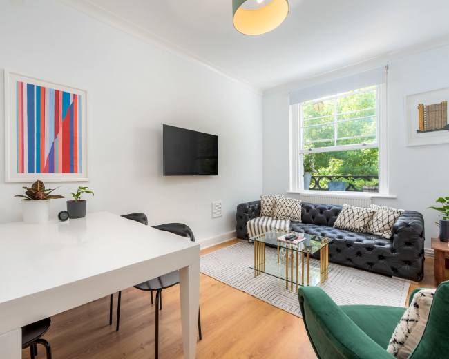 A modern living space features a black tufted sofa, a white dining table with black chairs, a wall-mounted television, and large windows letting in natural light.