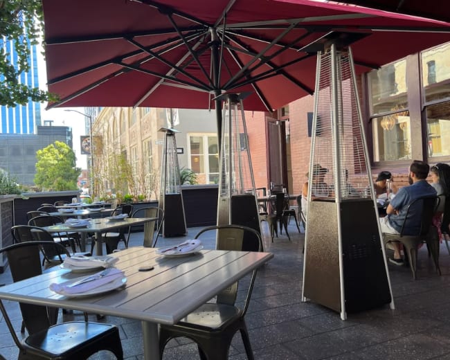 A patio dining area features outdoor tables with umbrellas and heat lamps, surrounded by greenery and a building in the background.