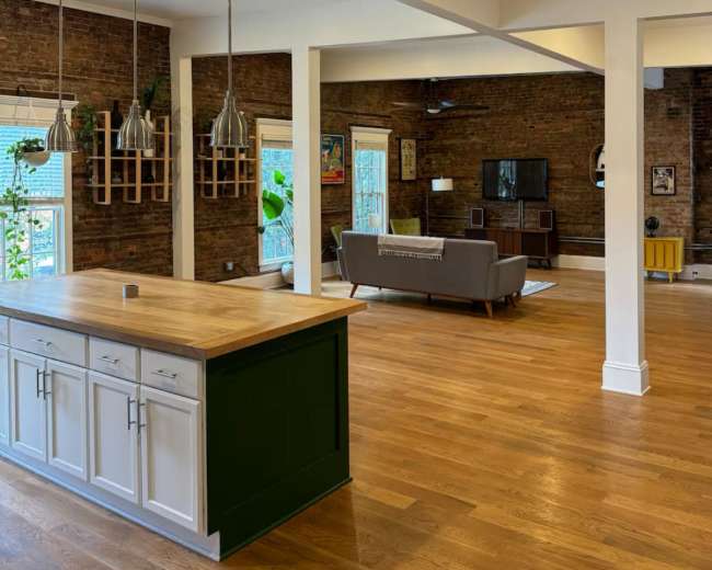 The image shows a spacious living area with brick walls, wooden flooring, and a kitchen island with a green base, leading to a cozy seating area.