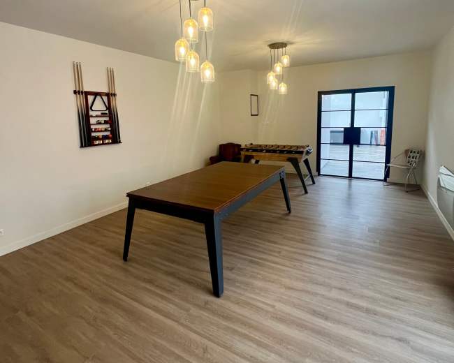 The image shows a spacious room with a large table, several light fixtures hanging from the ceiling, and a set of windows at one end.