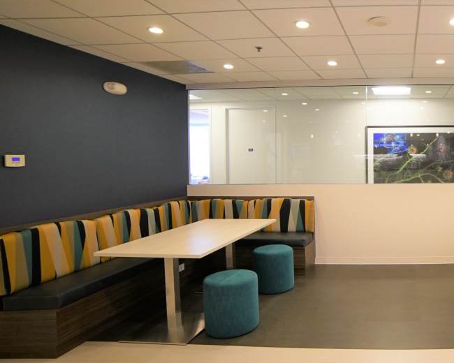 The image shows a modern seating area with two tables and bench seating, surrounded by blue and yellow striped cushions, along with teal stools in front of a glass wall.