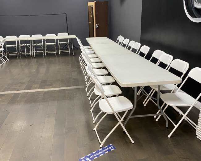 A long light-colored table is flanked by white folding chairs in a room with dark walls and a television mounted on one side.