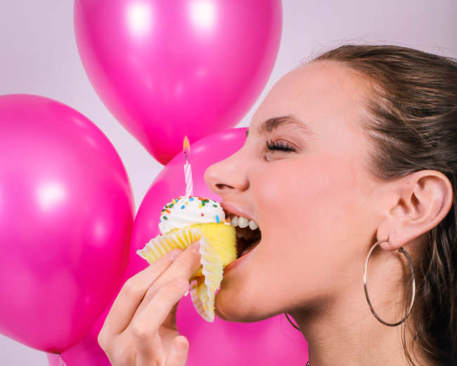 A woman with long hair is taking a bite of a cupcake while standing in front of pink balloons.