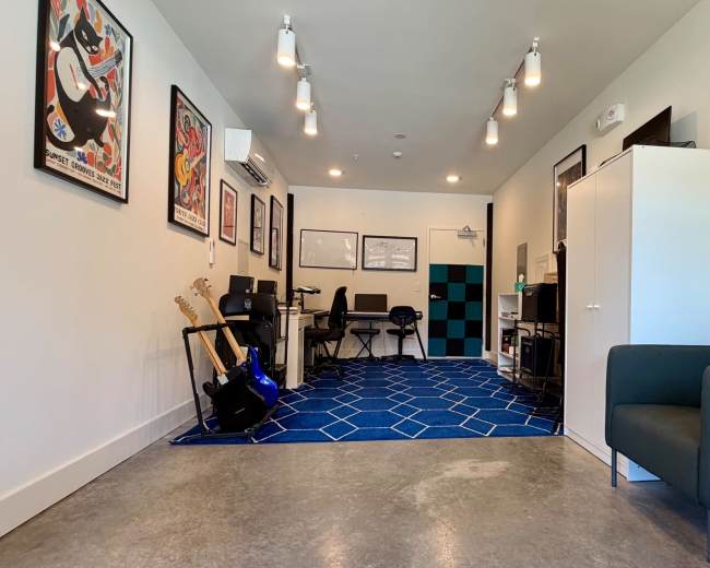 The image shows a modern music practice room featuring guitars, office chairs, a desk, and colorful wall art.