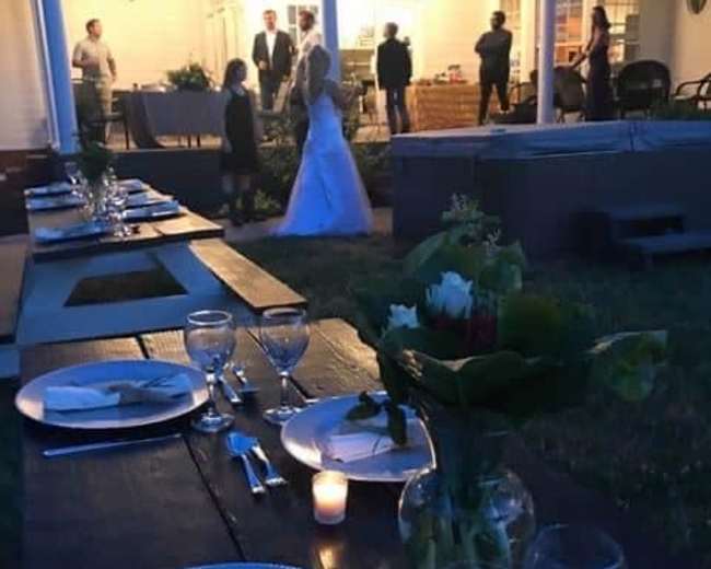 A beautifully arranged outdoor dining table is set with plates and glasses, while a wedding ceremony takes place in the background.