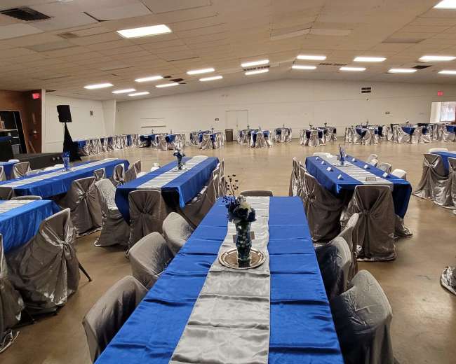 A spacious event hall is set up with long tables covered in blue tablecloths and accented with gray chair covers, arranged neatly for a gathering.