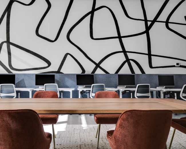 A large wooden table is surrounded by upholstered chairs in a modern office space with multiple computer workstations against a wall featuring an abstract black line mural.