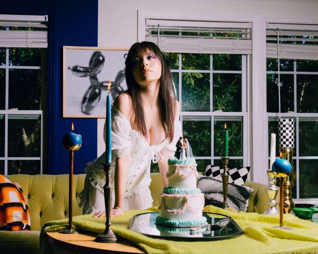 A person stands beside a three-tiered cake on a table adorned with candles and colorful decor in a well-lit room with large windows.