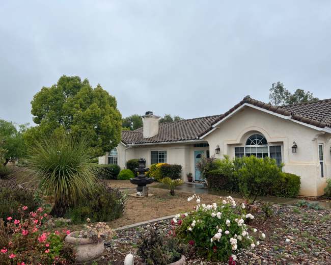 The image shows a single-story house with a tiled roof, surrounded by a landscaped yard featuring various shrubs and flower beds.