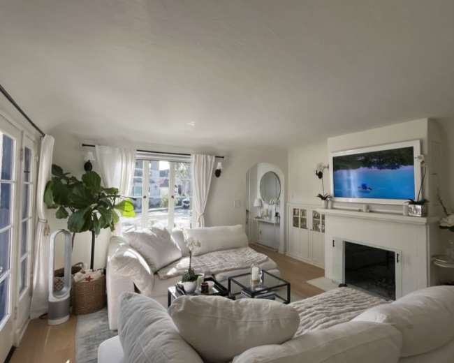 The living room features two large white sofas, a black coffee table, a flat-screen TV mounted above a white fireplace, and large windows with sheer curtains, allowing natural light to enter.