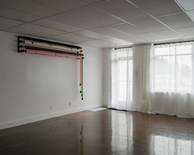 The image shows a spacious, empty room with a polished floor, large windows draped with sheer curtains, and a wall-mounted rack containing rolls of colored material.