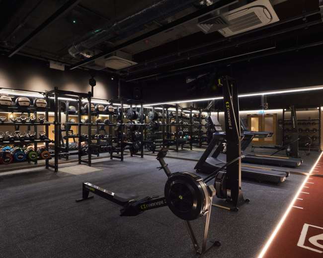 The image shows an indoor gym with various exercise equipment, including free weights, a rowing machine, and a treadmill, organized in a well-lit space.