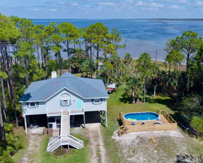 A two-story house with a gray roof is situated near a body of water, surrounded by tall pine trees and featuring a small above-ground pool in the yard.