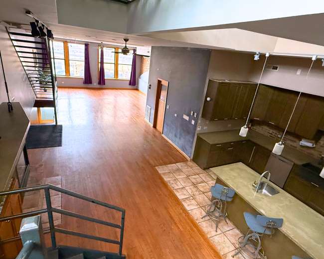 The image shows a spacious, open-concept loft with hardwood flooring, large windows, and a kitchen area featuring modern appliances and seating at a bar.