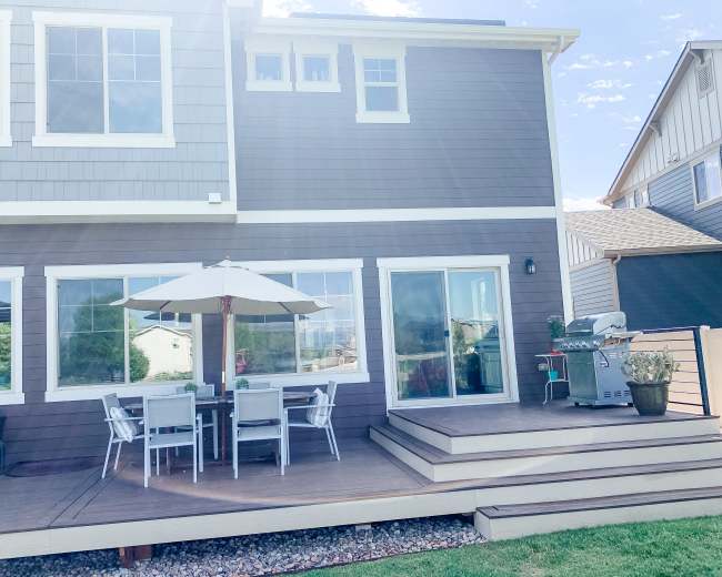 A patio area with a table and chairs under an umbrella, adjacent to a two-story house with large windows.