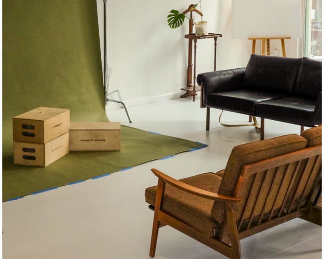 The image shows a minimalist studio setup with a green backdrop, two wooden boxes, a black leather sofa, and a wooden chair, all arranged on a white floor.