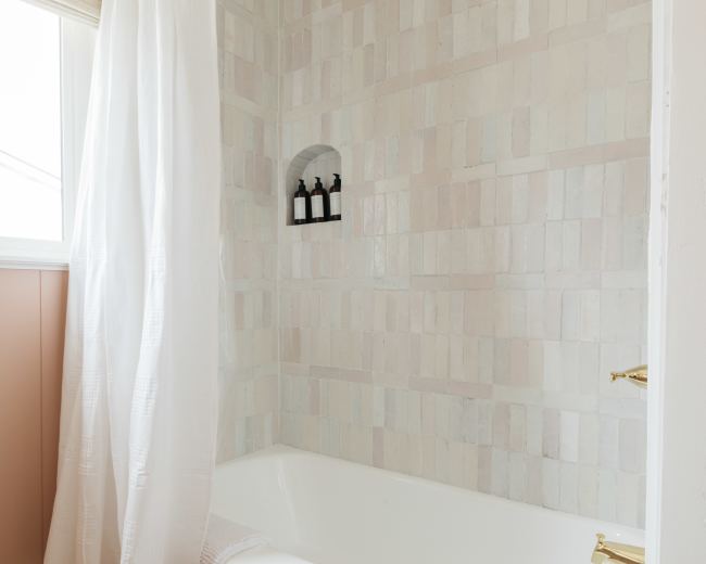 The image shows a bathroom with a white bathtub, beige hexagonal tiled floor, and a shower area enclosed by a white curtain, with light-colored, patterned tiled walls.